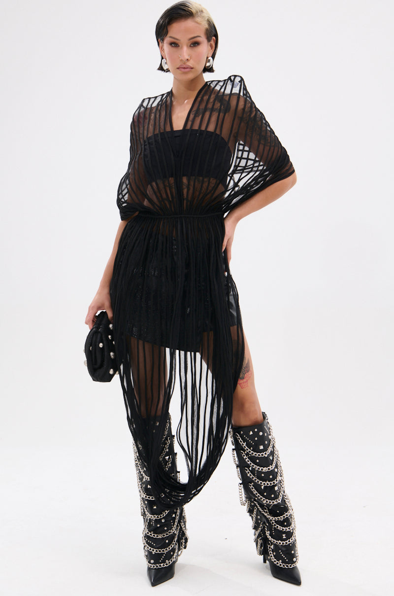 A person stands confidently against a plain white background, wearing the RIPPLING SHEER HALTER TOP over a black outfit with studded knee-high boots, chains, and holding a small black handbag for an avant-garde look.