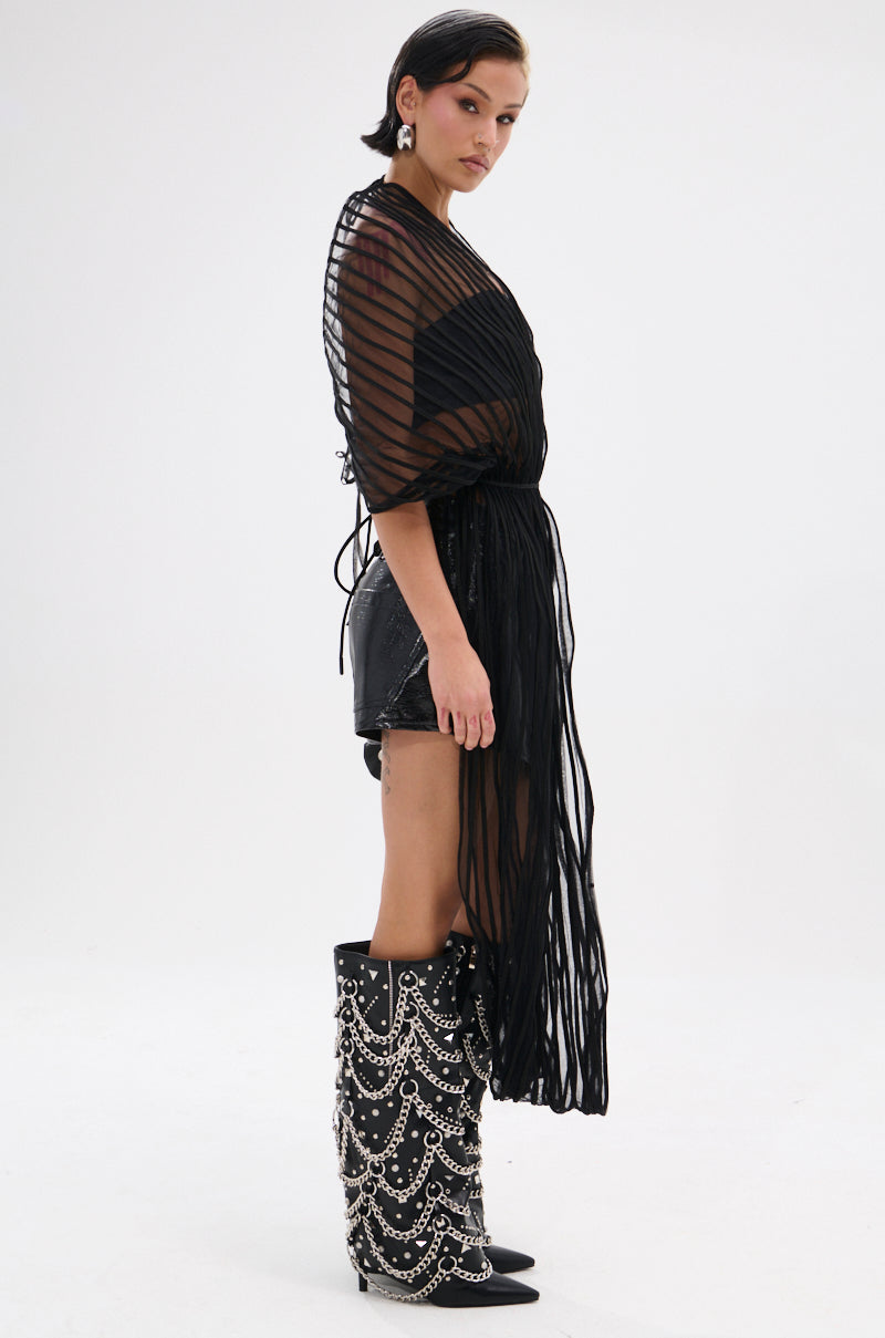 A person stands in profile against a white background, wearing the RIPPLING SHEER HALTER TOP over a black outfit and tall black boots with silver chains. The top features sheer, striped fabric with structured mesh details.