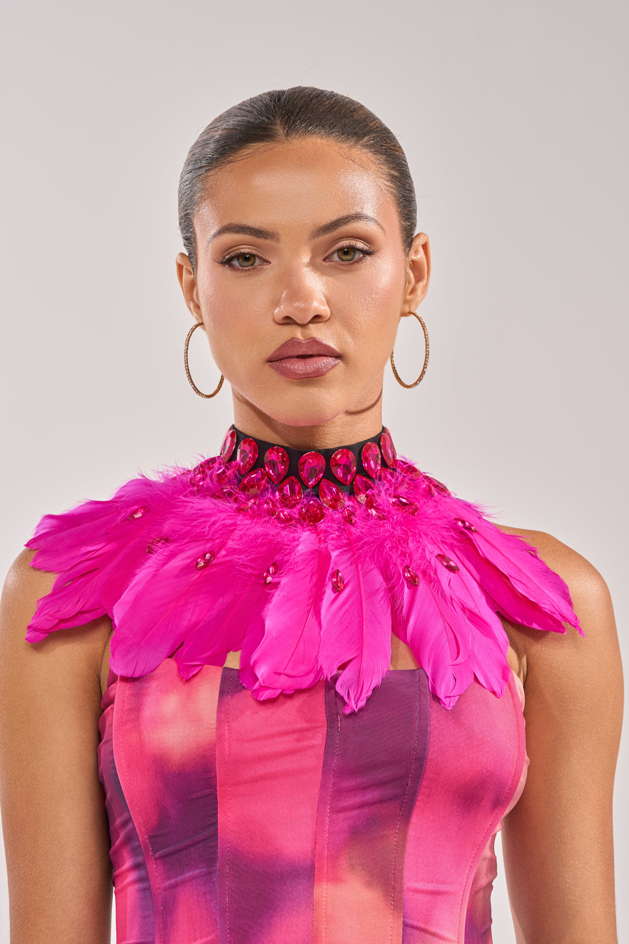 A woman with slicked-back hair wears large hoop earrings and the TAKE MY PIC FEATHER COLLAR NECKLACE, featuring vibrant pink feathers and red gems, styled with a colorful strapless top against a plain light background.