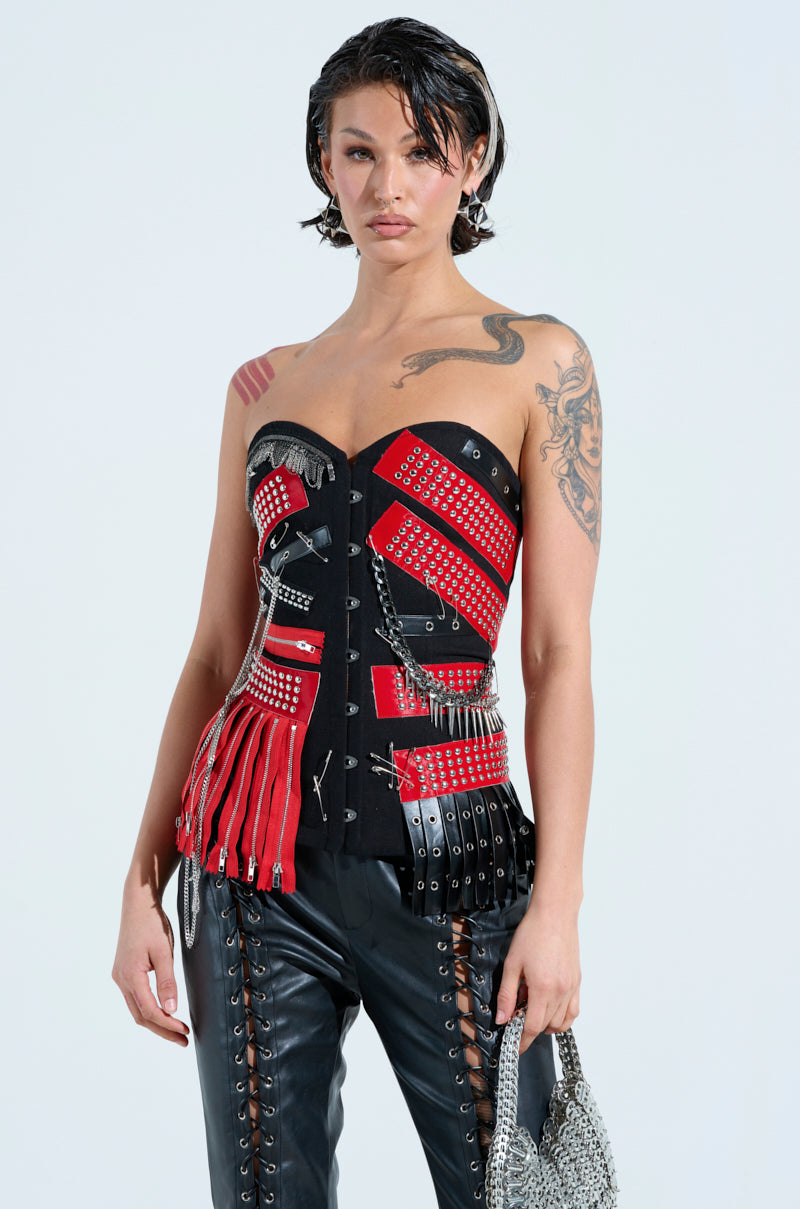 A rock star with slicked-back hair sports the DEMI CHAIN CORSET, a strapless top with red and black panels, studs, and fringe. Paired with black leather lace-up pants, a small metallic bag, and visible arm tattoos for an edgy look.