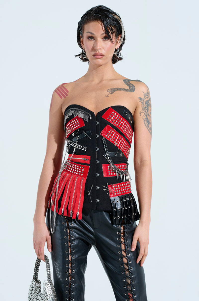 With wet hair exuding rock star vibes, they wear the DEMI CHAIN CORSET—a strapless black and red number with studs and zippers—paired with side-laced black leather pants, carrying a shiny silver bag and showcasing tattoos on their arms and shoulder.