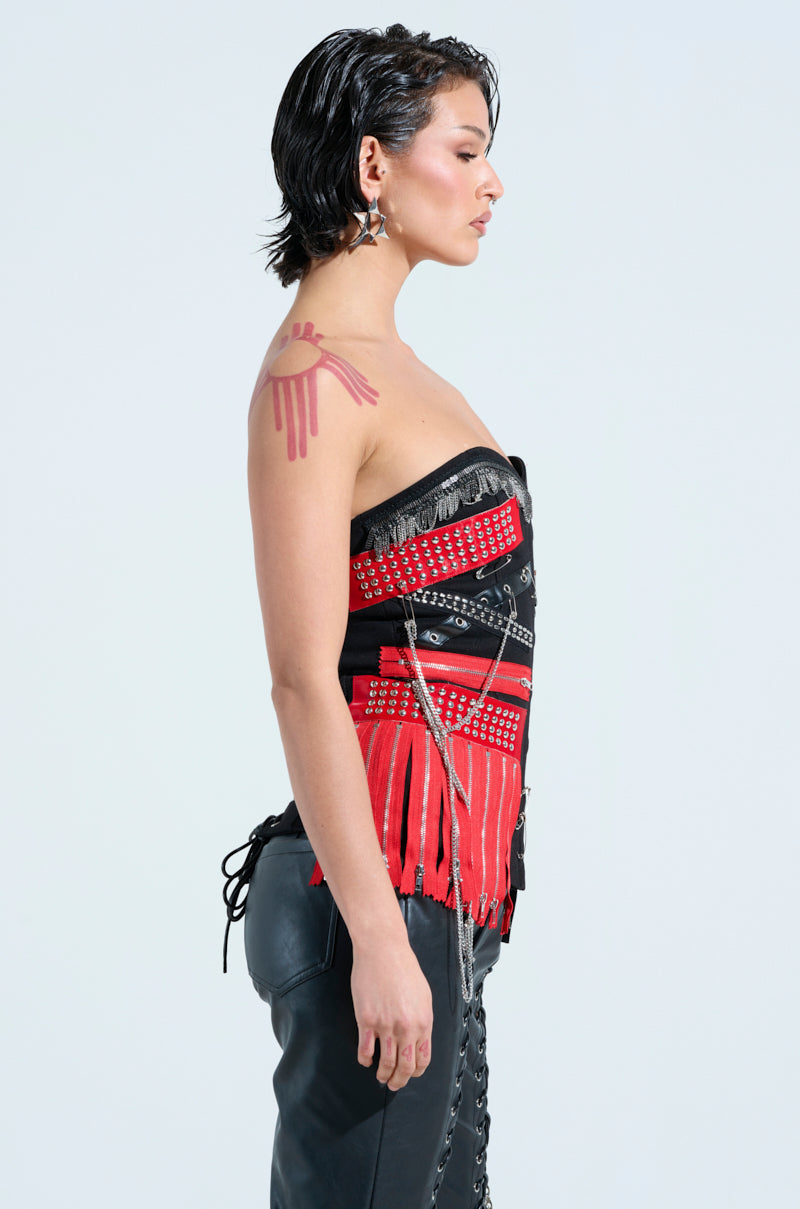A person with short dark hair stands in profile against a plain background, exuding rock star vibes. They are wearing a DEMI CHAIN CORSET with fringe and metal accents, paired with black lace-up pants. A striking red tattoo adorns their left shoulder.