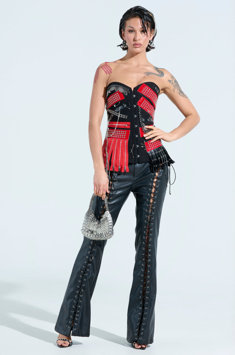A person poses in the DEMI CHAIN CORSET, a studded, rock-star inspired red and black strapless top with fringe. It's paired with black leather pants featuring lace-up sides. They hold a studded silver handbag and wear sandals, their hair styled in a dark, wet look against a white background.