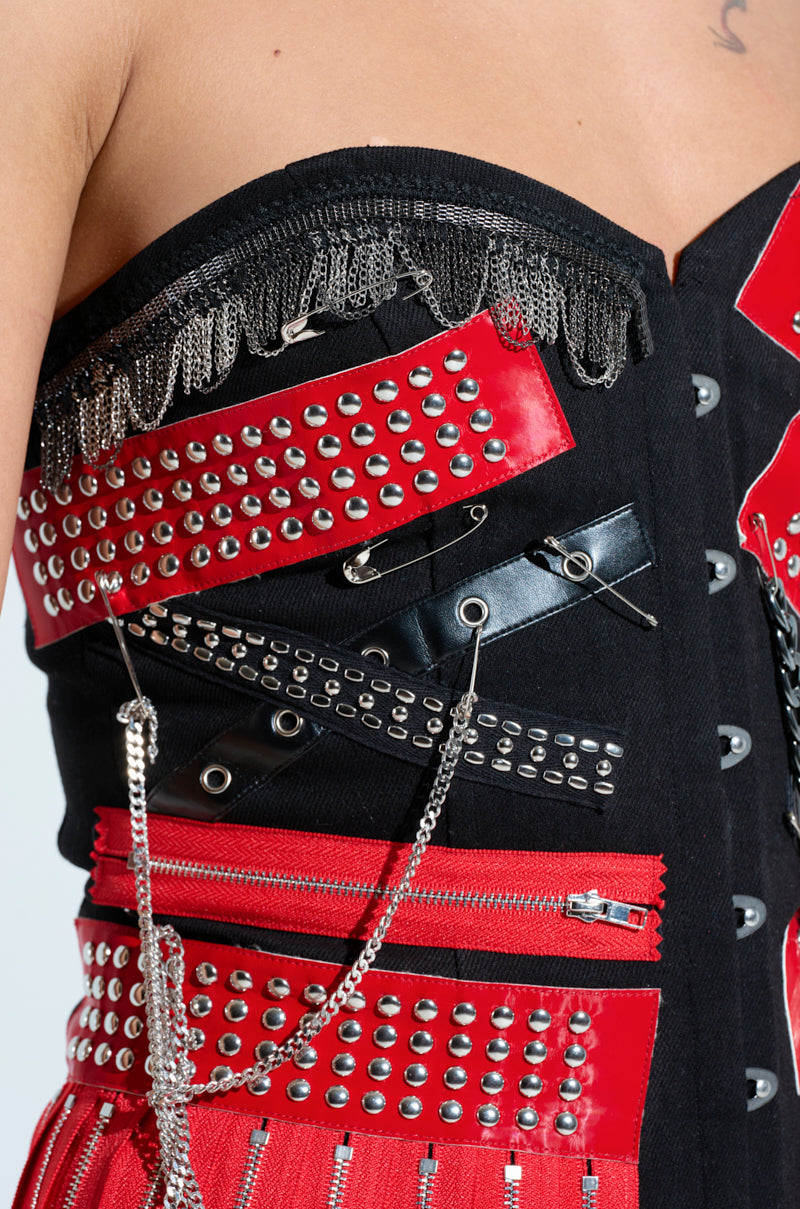 A close-up of the DEMI CHAIN CORSET shows a rock star style with red and black fabric, studded embellishments, safety pins, dangling chains, and metal fringe detailing along the top edge for an edgy flair.