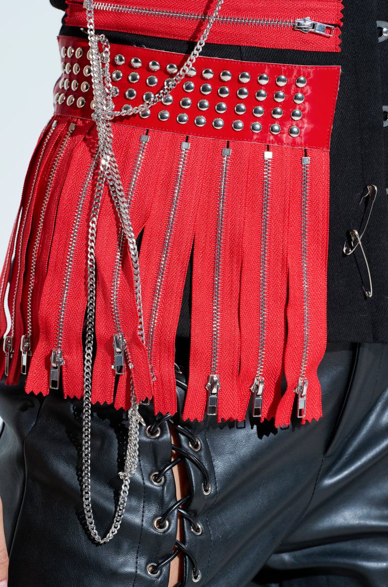 Close-up of a fashion-forward ensemble featuring a red fringed belt with studs and silver chains over shiny black lace-up leather pants. Paired with the DEMI CHAIN CORSET, this look combines edgy and modern elements, evoking a rock star vibe with bold colors and textures.