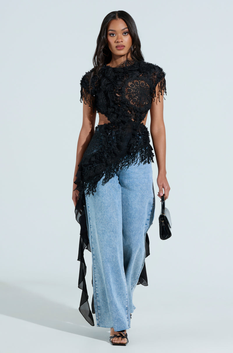 Wearing the THINK ABOUT ME TOP IN BLACK with fringe and a small black handbag, the person walks confidently in light blue wide-legged jeans. The metallic back zip of the top glints against a light background.