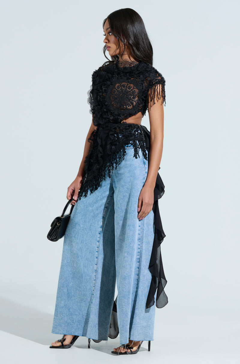 A woman stands in profile wearing the THINK ABOUT ME TOP IN BLACK and light blue wide-leg jeans. She's holding a small black purse, wearing open-toed heels, and has her long hair loose with sheer mesh draping from her waist.