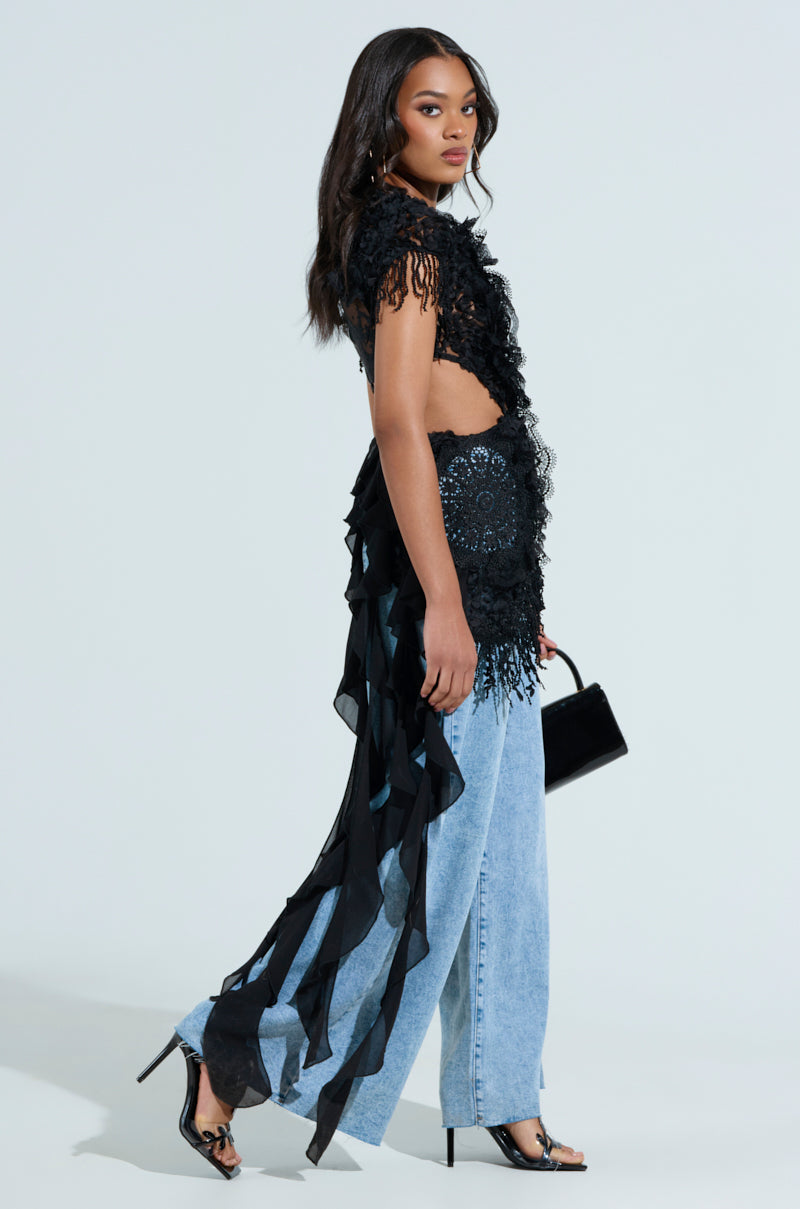 A woman wears the THINK ABOUT ME TOP IN BLACK, featuring floral lace ruffles and sheer mesh panels, paired with light blue wide-leg jeans. She holds a small black purse and dons black high-heeled shoes against a simple gray backdrop.