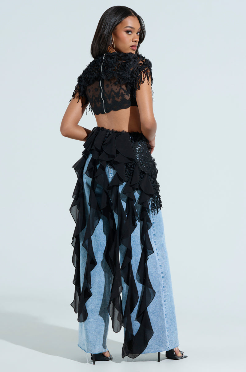 A woman poses with her back to the camera in a chic THINK ABOUT ME TOP IN BLACK, featuring sheer mesh and flowing ruffles, paired with light blue wide-leg jeans. Her long hair cascades freely on a plain background, while a metallic back zip adds an edgy touch.