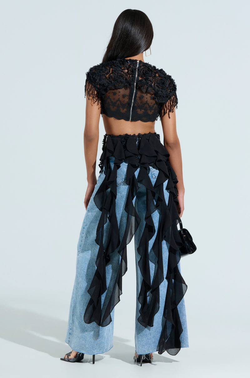 A woman with long, straight hair faces away, showcasing the THINK ABOUT ME TOP IN BLACK featuring floral lace and ruffles. She pairs it with wide-leg denim jeans, black heels, and holds a small black handbag against a neutral background.