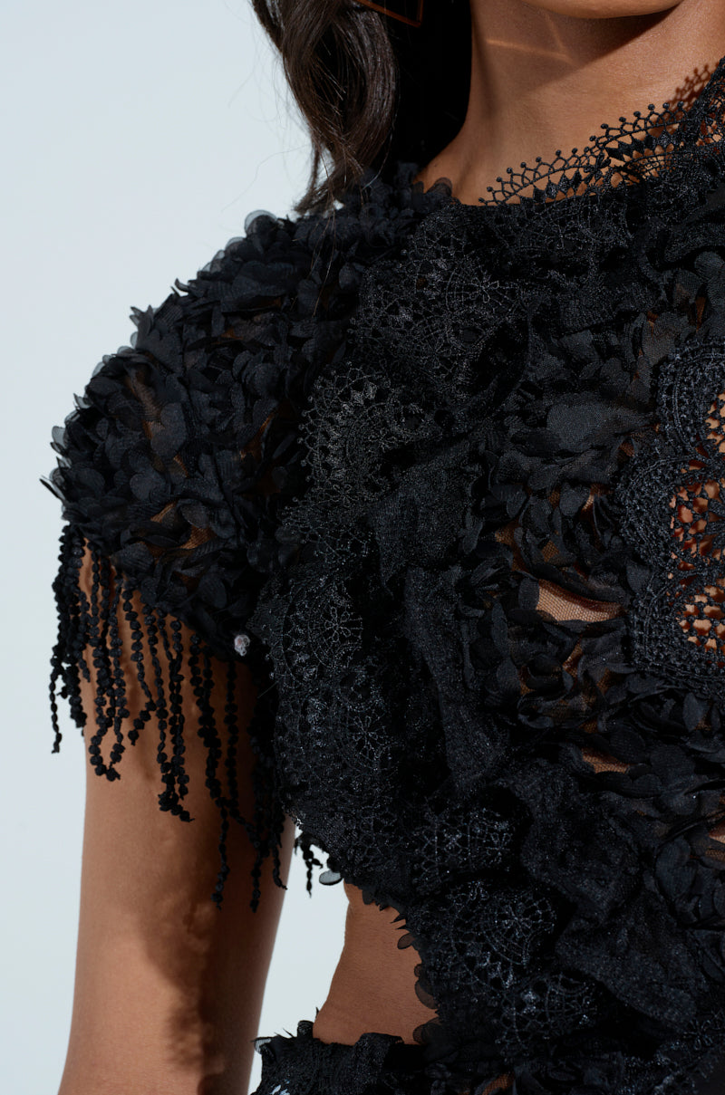 Close-up of a person wearing the THINK ABOUT ME TOP IN BLACK, featuring intricate floral lace patterns and fringe accents. The sheer mesh and metallic back zip add varied textures, creating an elegant look against a light background.