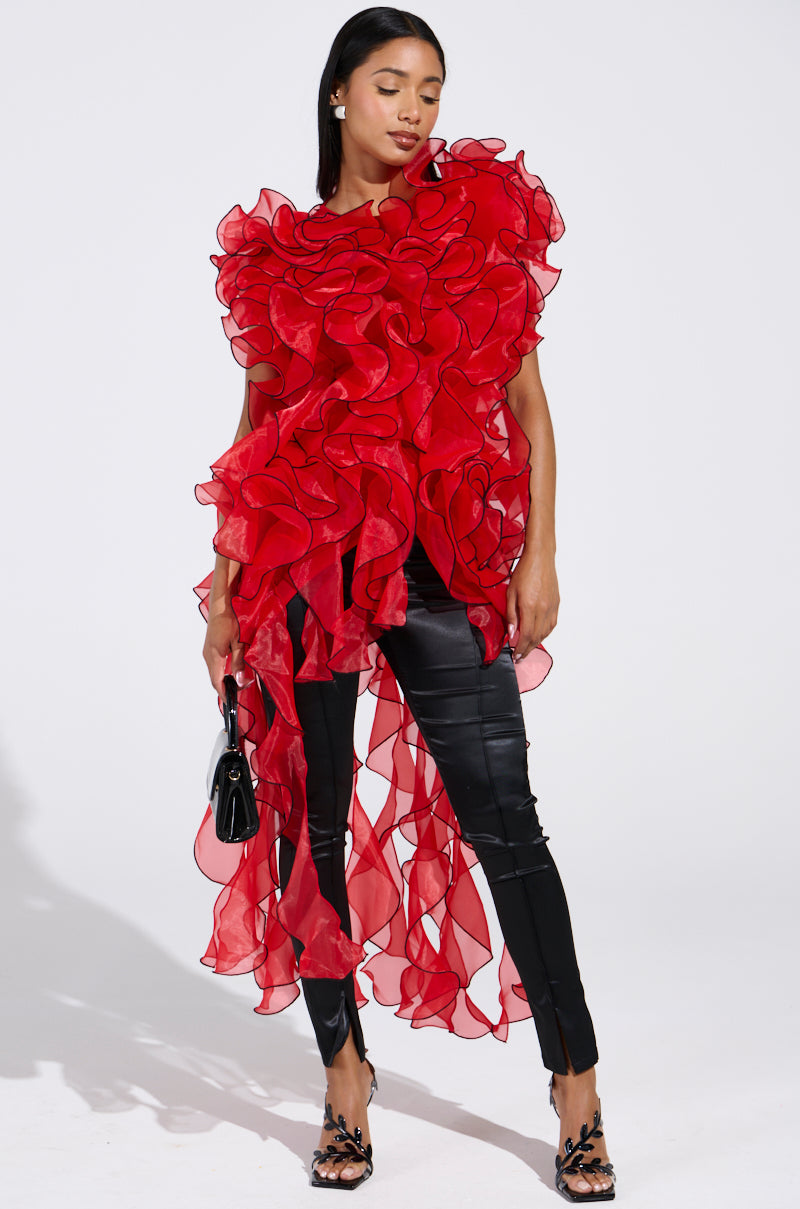 A woman models the LOVER RUFFLE BLOUSE, featuring dramatic red asymmetrical ruffles and sheer organza layers, styled with black leather pants, black heels, and a small black handbag against a white background.