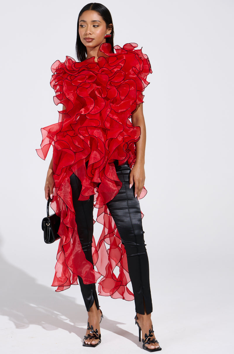A woman poses against a white background wearing the LOVER RUFFLE BLOUSE, featuring bold sheer organza with dramatic asymmetrical ruffles. She pairs it with black fitted pants, strappy heels, and carries a small black handbag.