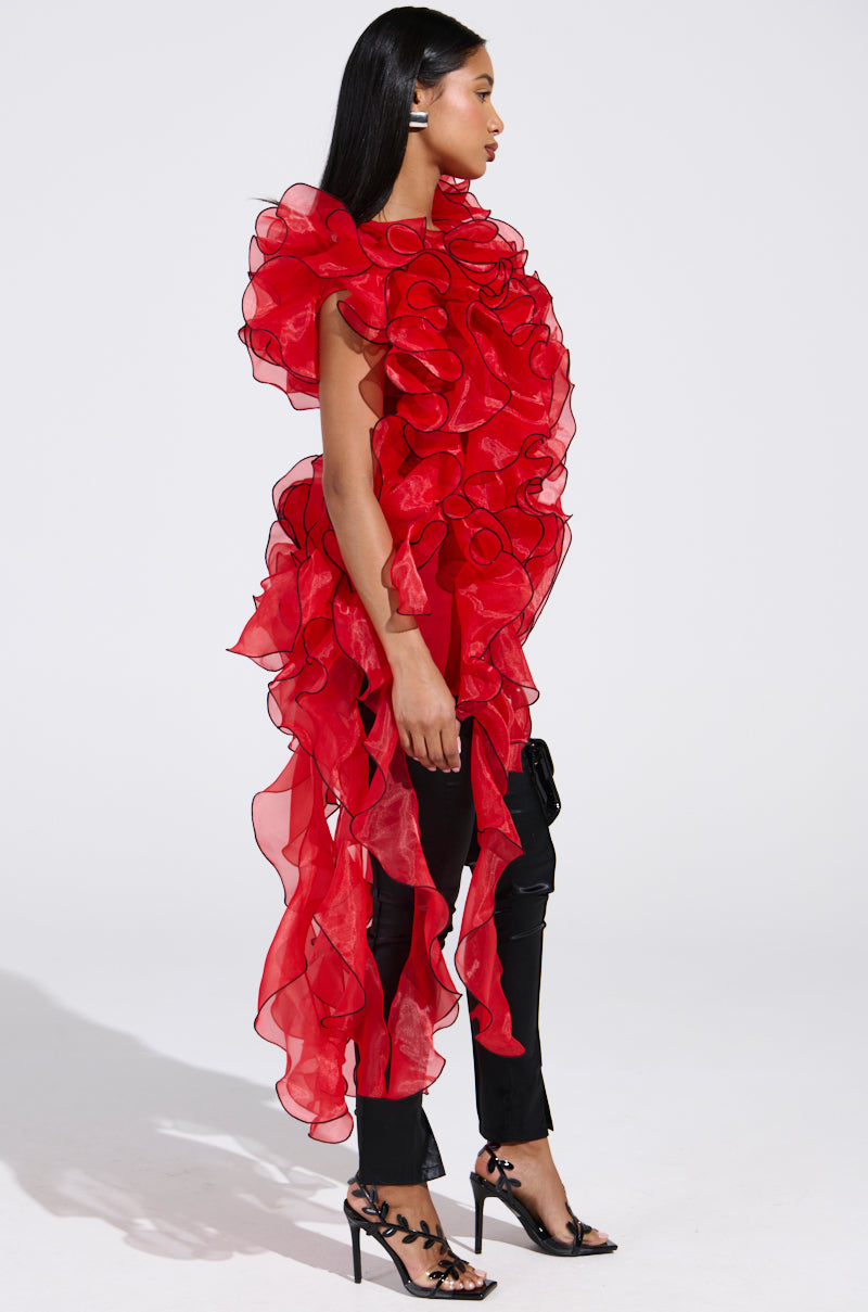 A woman stands sideways against a white background, wearing the LOVER RUFFLE BLOUSE in sheer organza with black trim, paired with slim black pants and high-heeled sandals. Her straight hair falls loosely past her shoulders.