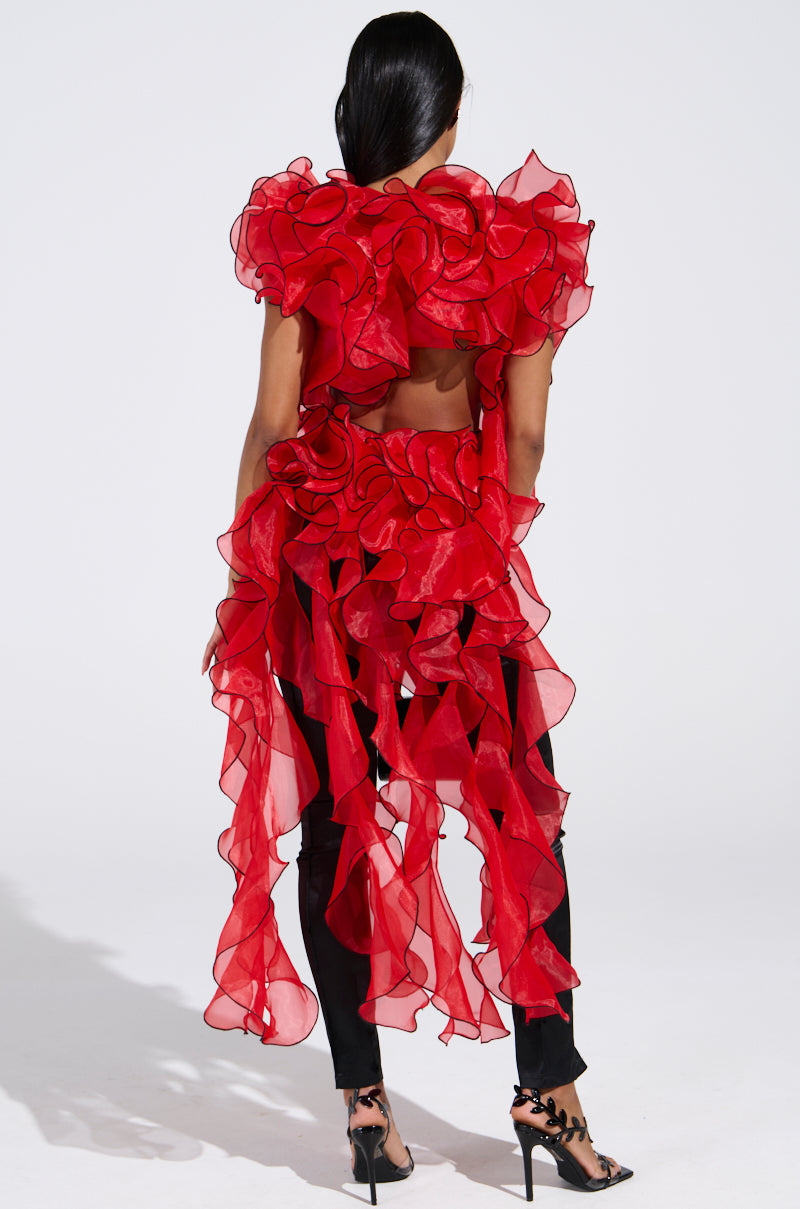 A person stands facing away, wearing the LOVER RUFFLE BLOUSE—a sheer red organza top with asymmetrical ruffles and long layers—paired with black pants, high-heeled sandals, and their long dark hair tied back.