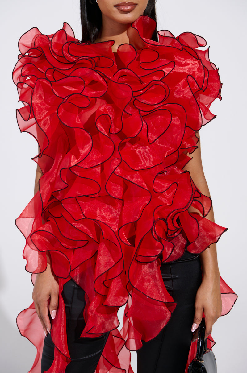 A woman wears the LOVER RUFFLE BLOUSE, featuring vibrant sheer organza with black edging and dramatic, asymmetrical layered ruffles, paired with black pants and a small black purse.