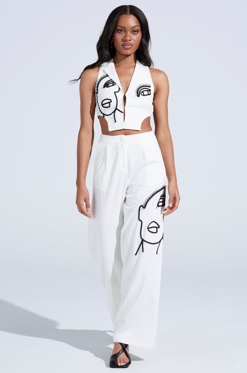 A woman confidently walks in a white crop top and MAYBE NEXT TIME TROUSER, featuring abstract black line art on the high-rise waist pants. She completes her look with black heels against a light neutral background.