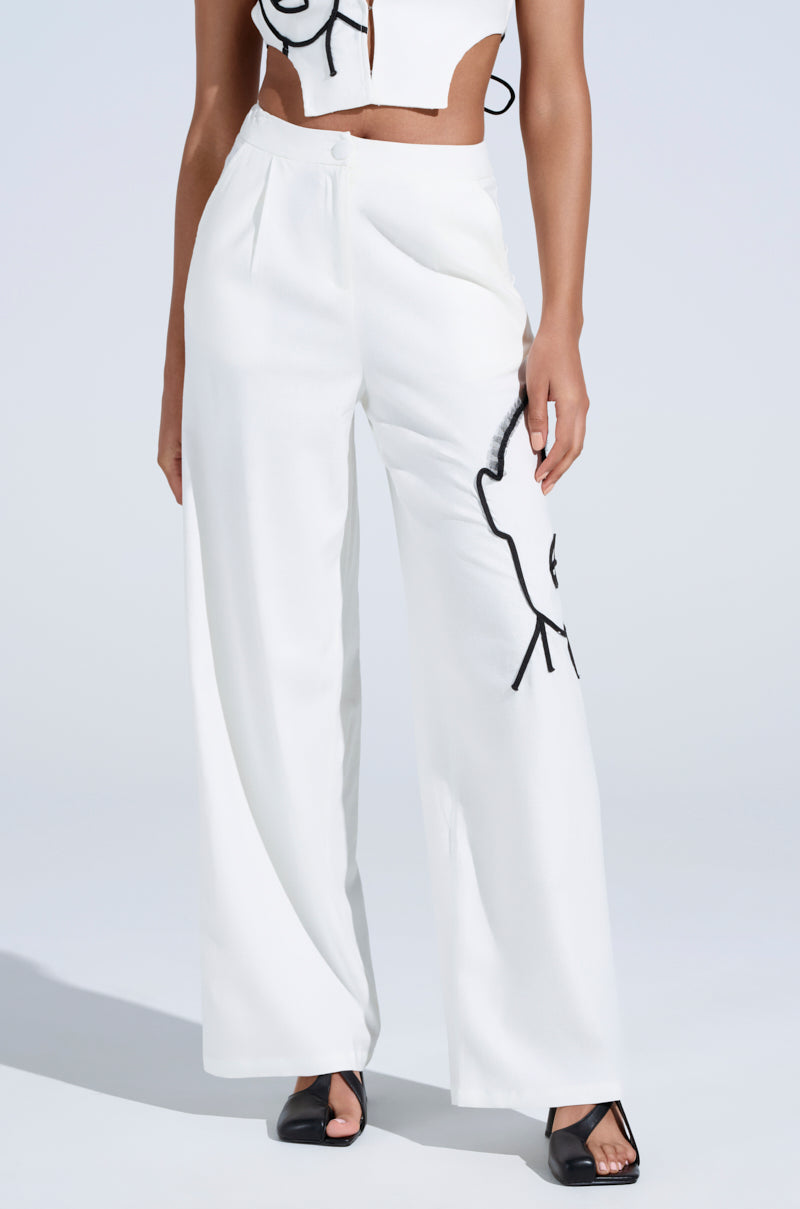 Wearing MAYBE NEXT TIME TROUSER, a person displays white wide-leg pants with a high-rise waist and abstract embroidery on one leg, paired with a matching white crop top. Black open-toe heels complement the outfit against a plain, light backdrop.