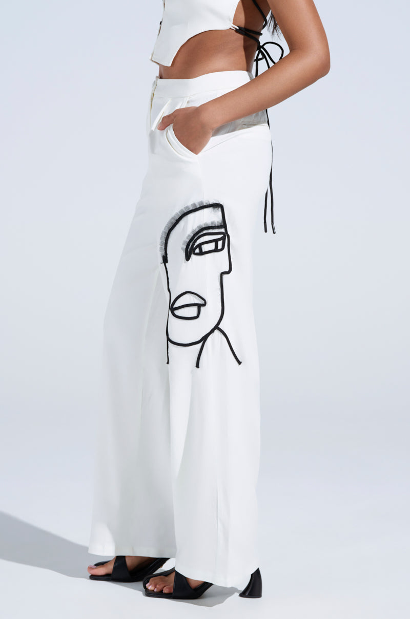 A person wears the MAYBE NEXT TIME TROUSER, featuring a white wide-leg design with an abstract embroidered face on one side and a high-rise waist, paired with a white crop top. They stand against a light background, hand in pocket.