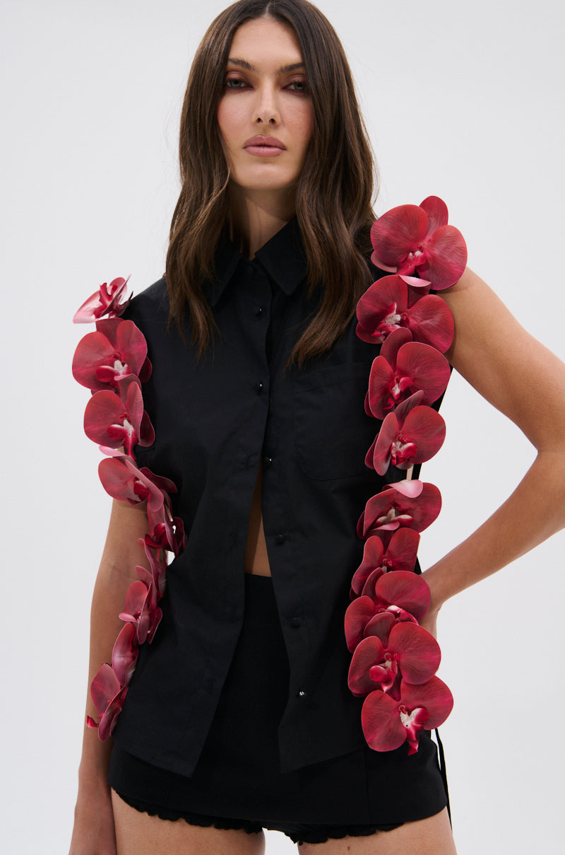 A woman models the ROSE PETALS BUTTON DOWN—a sleeveless black poplin cotton top with red orchid accents—paired with black shorts against a plain light background.