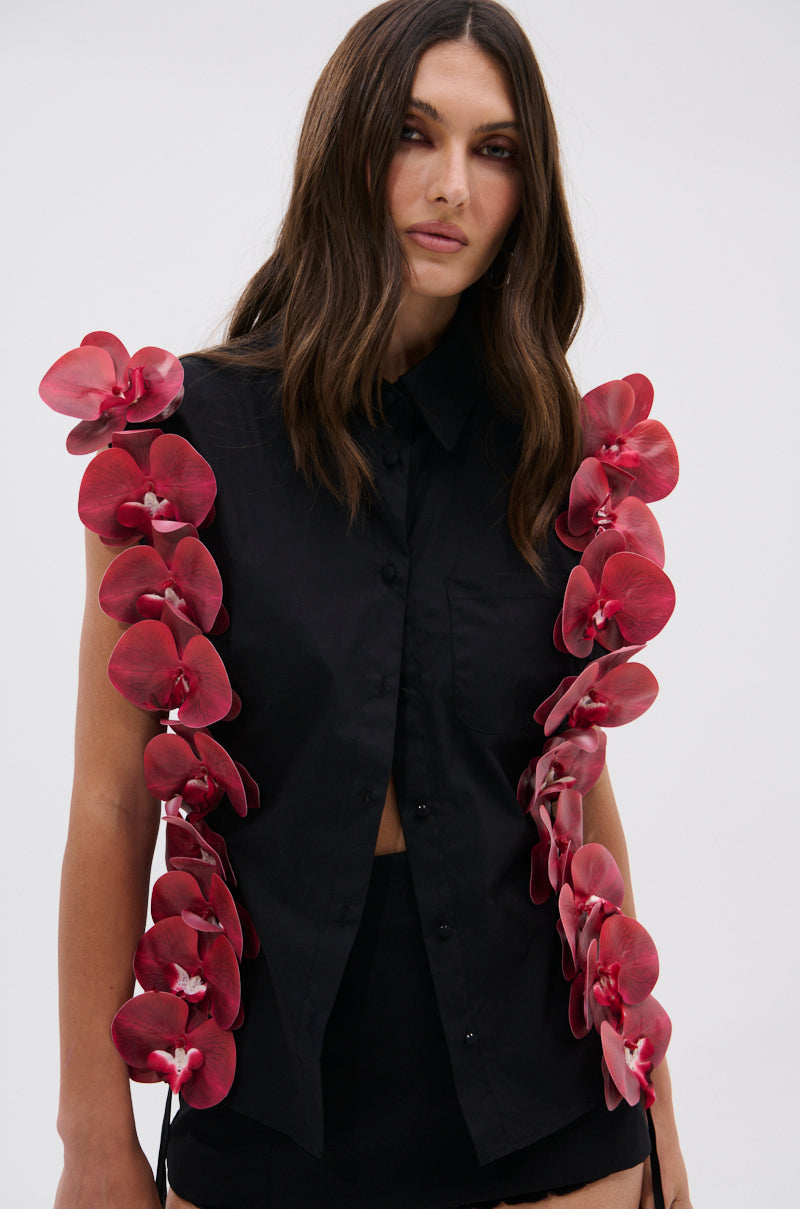 A woman stands against a plain light background, facing the camera with a neutral expression. She wears the ROSE PETALS BUTTON DOWN—a sleeveless black top featuring red orchid-like floral designs. Her long brown hair is worn loose.
