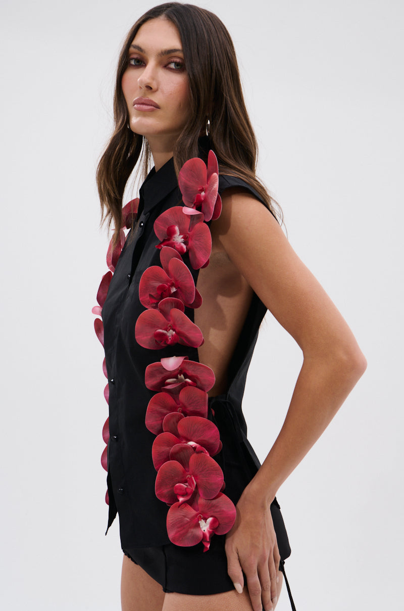 A woman with long, wavy hair and a neutral expression poses against a plain light background wearing the ROSE PETALS BUTTON DOWN, a sleeveless black poplin cotton top with 3D floral appliques along the front and side.