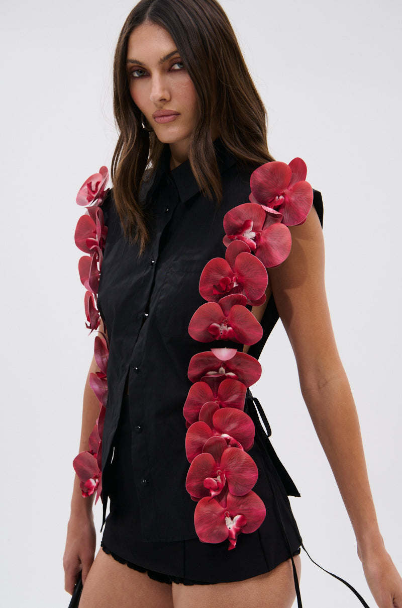 A woman wears the ROSE PETALS BUTTON DOWN, a sleeveless black poplin cotton top with large red 3D floral appliques on both sides, paired with matching black shorts, standing against a plain light background.