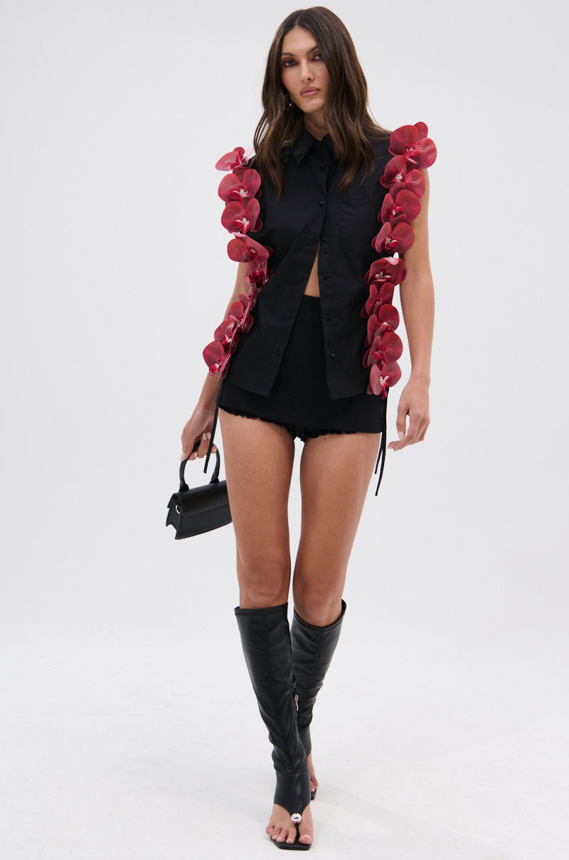 A woman stands against a plain white background wearing the ROSE PETALS BUTTON DOWN, a black sleeveless top with bold 3D floral appliques, paired with black shorts, knee-high open-toe boots, and holding a small black handbag.