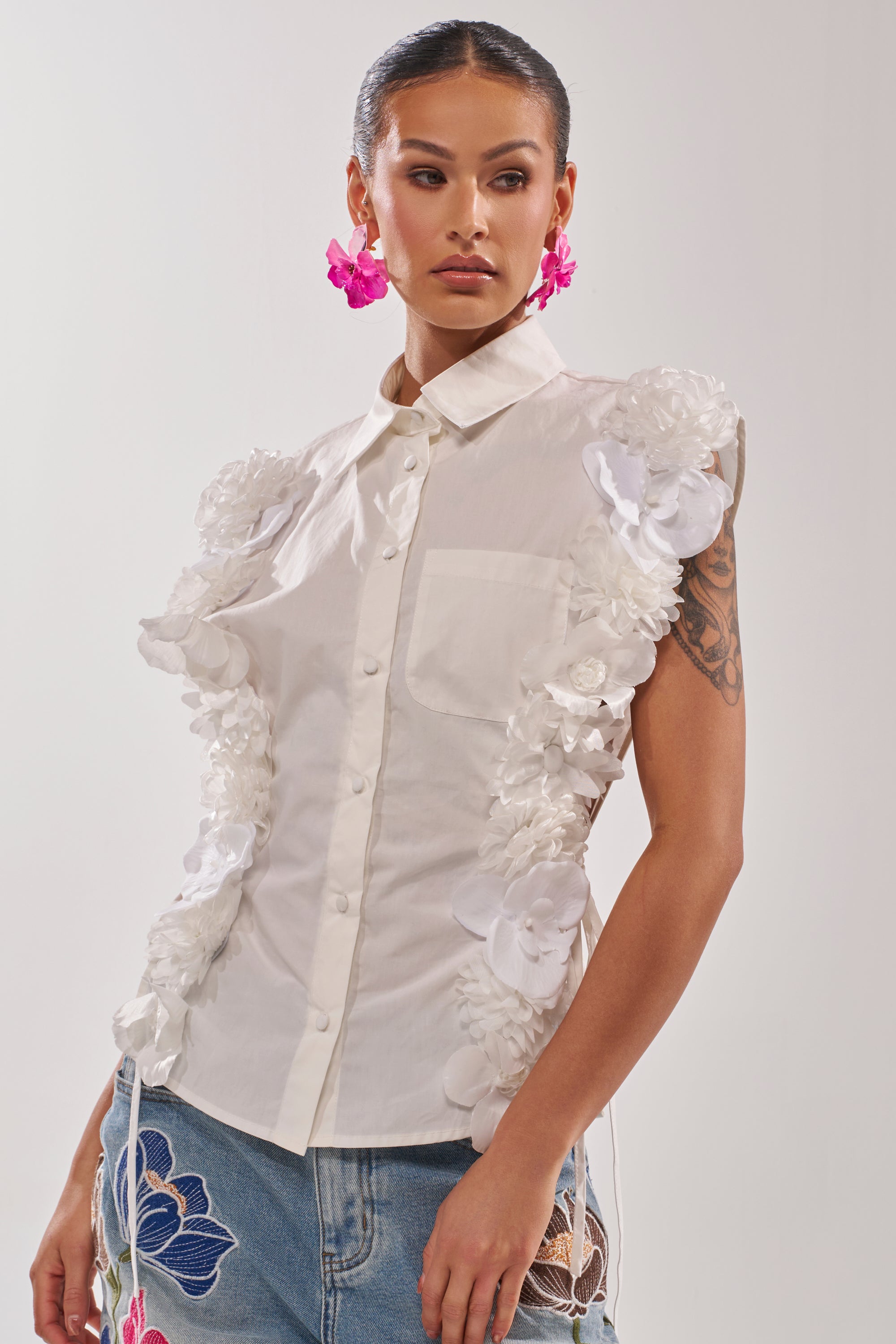 A woman wears the CRAZY FOR YOU BUTTON DOWN, a cotton blouse with short sleeves and white 3D floral appliqué on the sides, paired with blue embroidered jeans. She accessorizes with pink flower earrings and displays a visible arm tattoo. 0