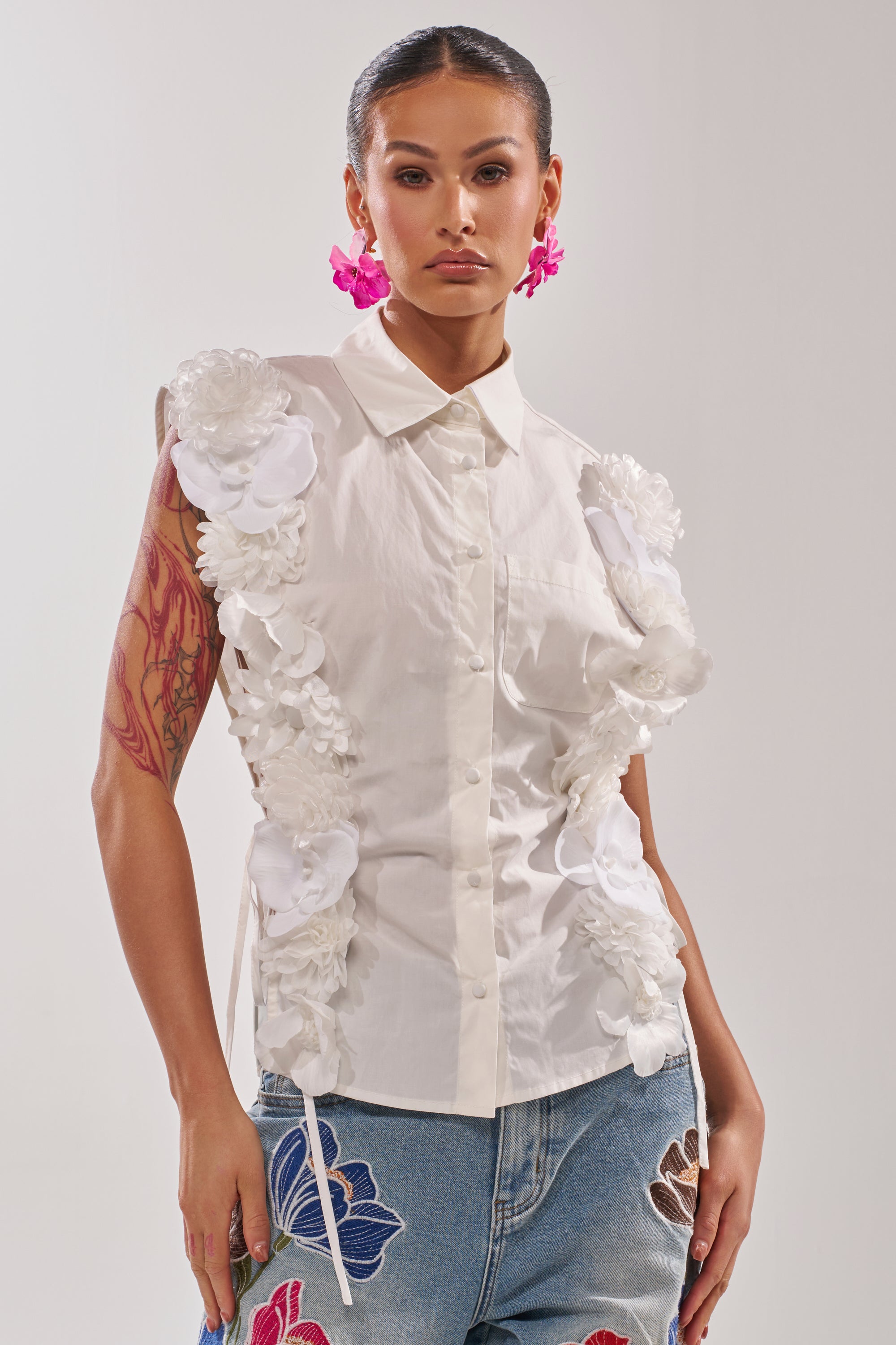 A woman wears the CRAZY FOR YOU BUTTON DOWN, a sleeveless cotton blouse with a collared neckline and white 3D floral appliqué. She pairs it with blue jeans featuring colorful floral embroidery, pink flower earrings, and slicked-back hair. 1