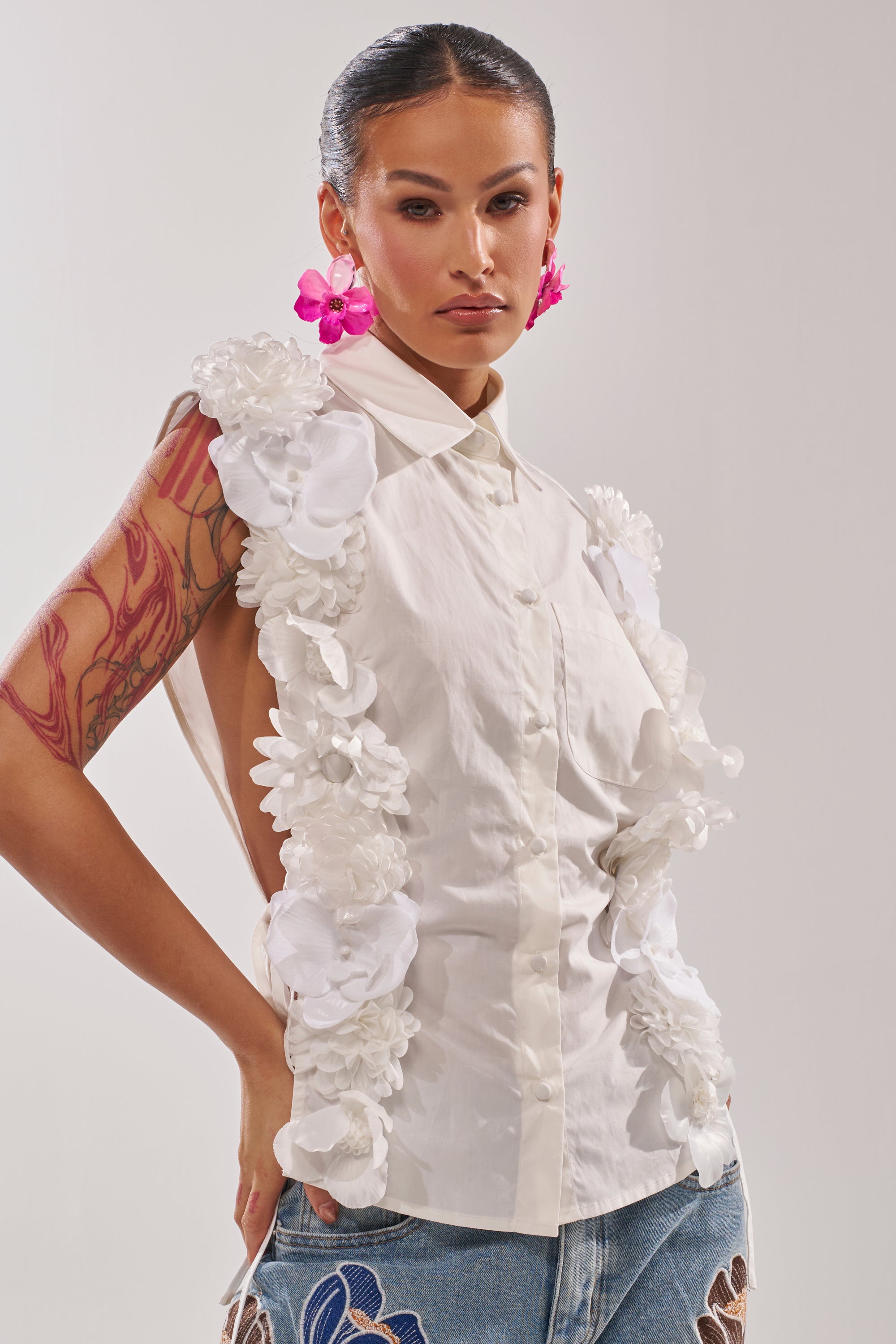 A woman wears the CRAZY FOR YOU BUTTON DOWN, a sleeveless cotton blouse with a collared neckline and 3D floral appliqué, styled with blue jeans featuring embroidered flowers, pink flower earrings, and a colorful arm tattoo against a plain background. 2
