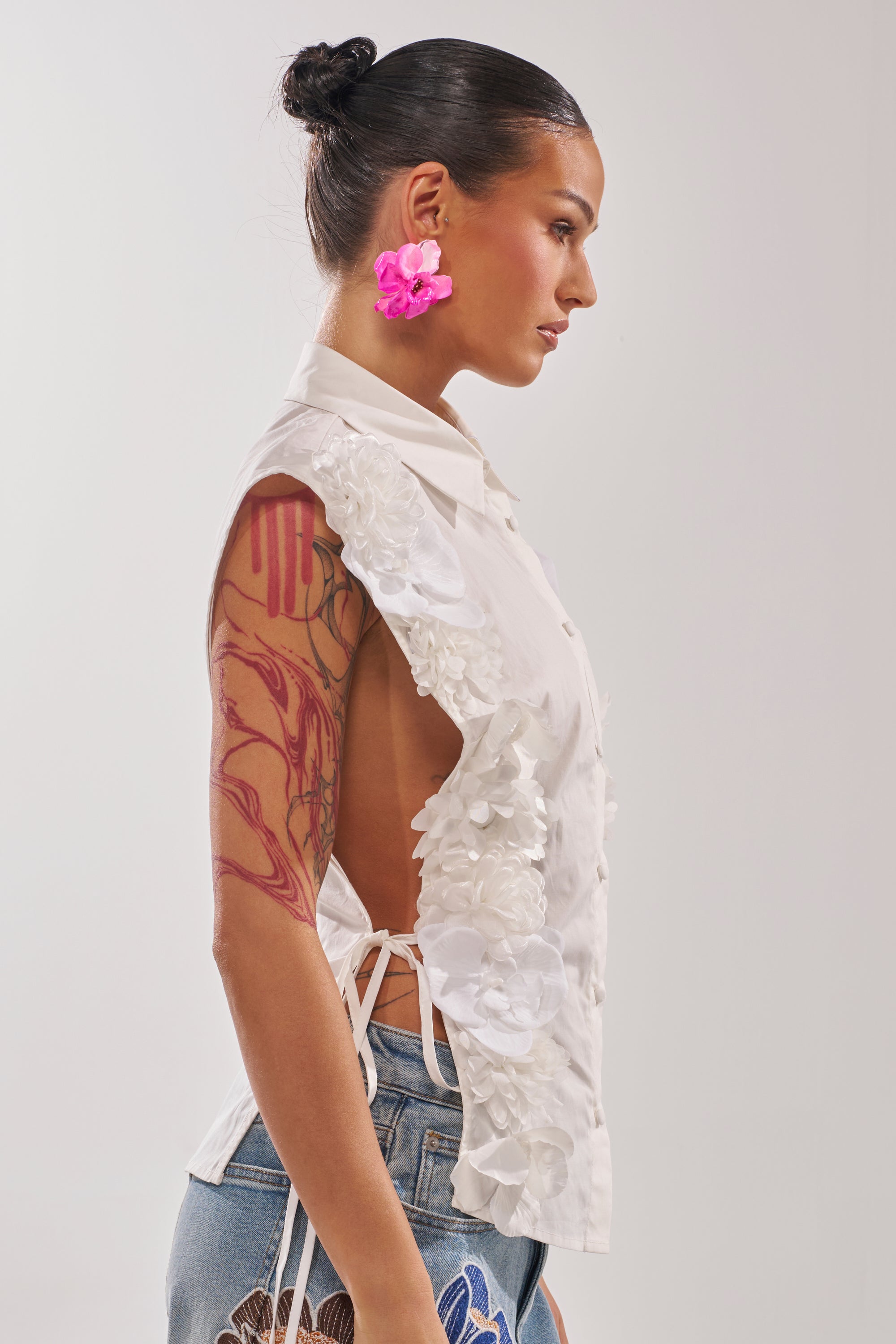 A woman with slicked-back hair and pink flower earrings wears the CRAZY FOR YOU BUTTON DOWN—a sleeveless cotton blouse with 3D floral appliqué and open sides—paired with denim jeans, showing off her vibrant tattoo sleeve. 3