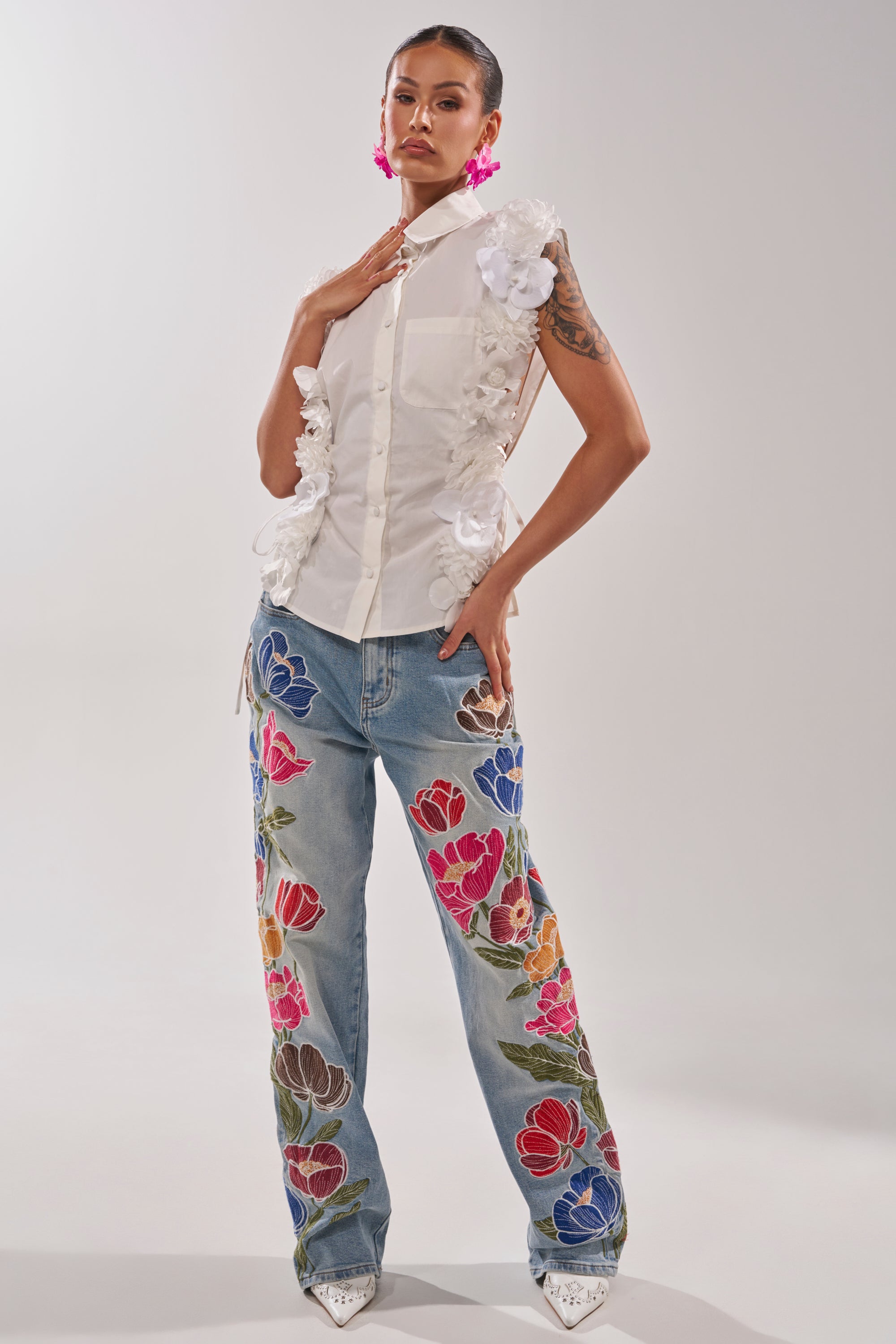 A woman wears the CRAZY FOR YOU BUTTON DOWN, a sleeveless cotton blouse with a collared neckline and 3D floral appliqué, paired with blue embroidered jeans, pink statement earrings, and pointed white shoes. 5