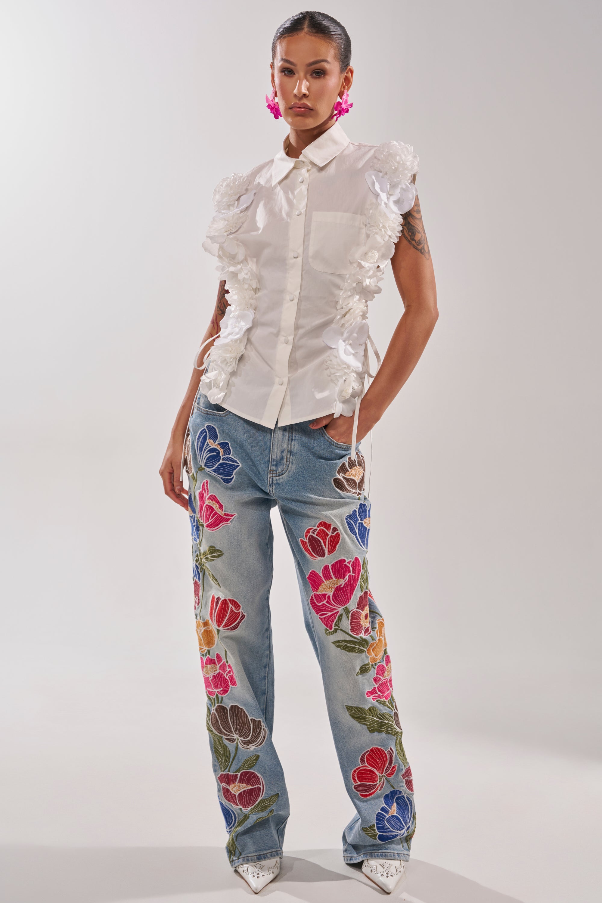 A woman stands confidently in the CRAZY FOR YOU BUTTON DOWN, a white collared cotton blouse, paired with blue jeans with bold floral embroidery, white pointed-toe shoes, and bright pink flower earrings. The background is plain and light. 6