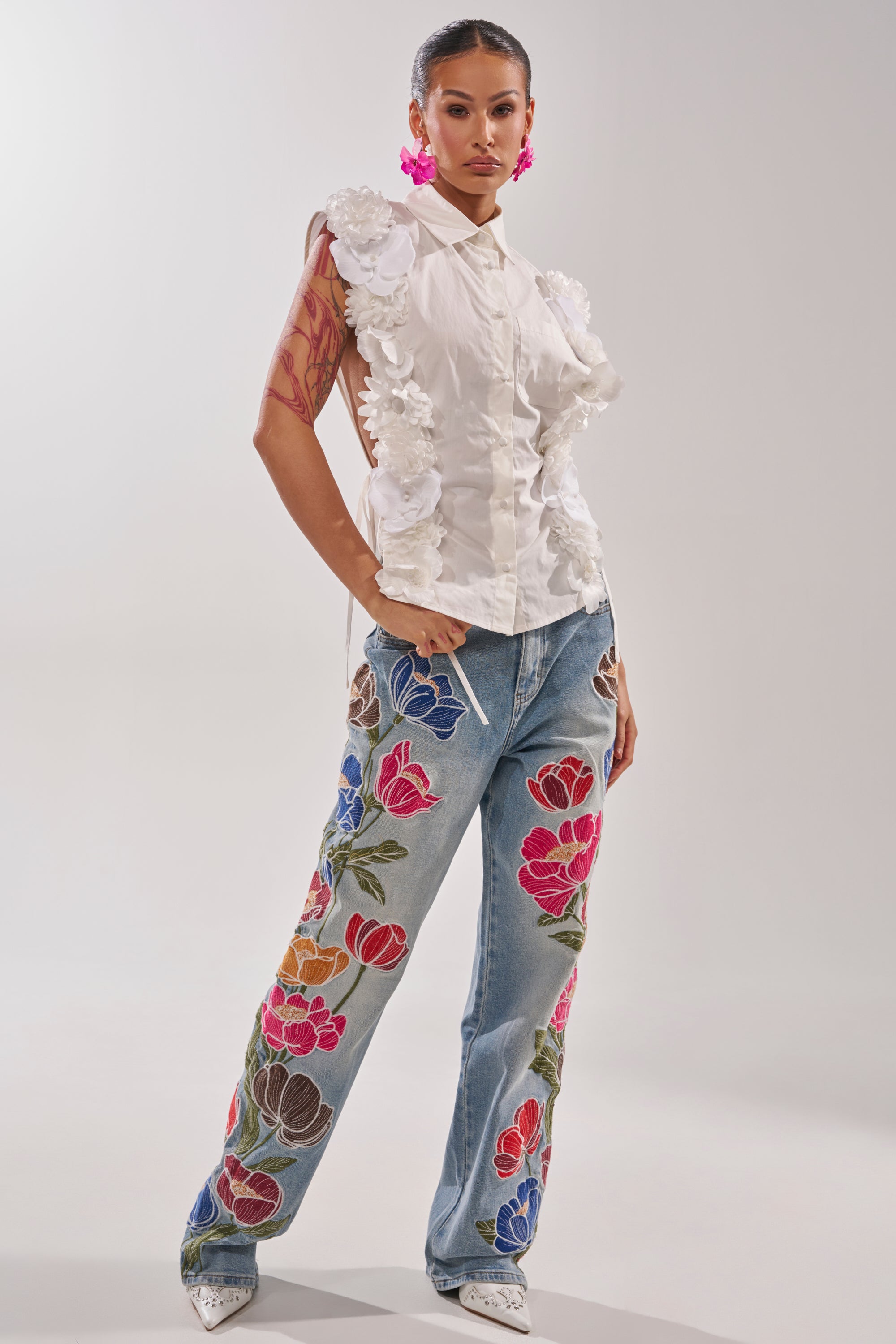 She stands confidently in the CRAZY FOR YOU BUTTON DOWN, a white blouse with 3D floral appliqué and collared neckline, blue jeans featuring colorful flower embroidery, white pointed shoes, pink floral earrings, and visible tattoos on her right arm. 7