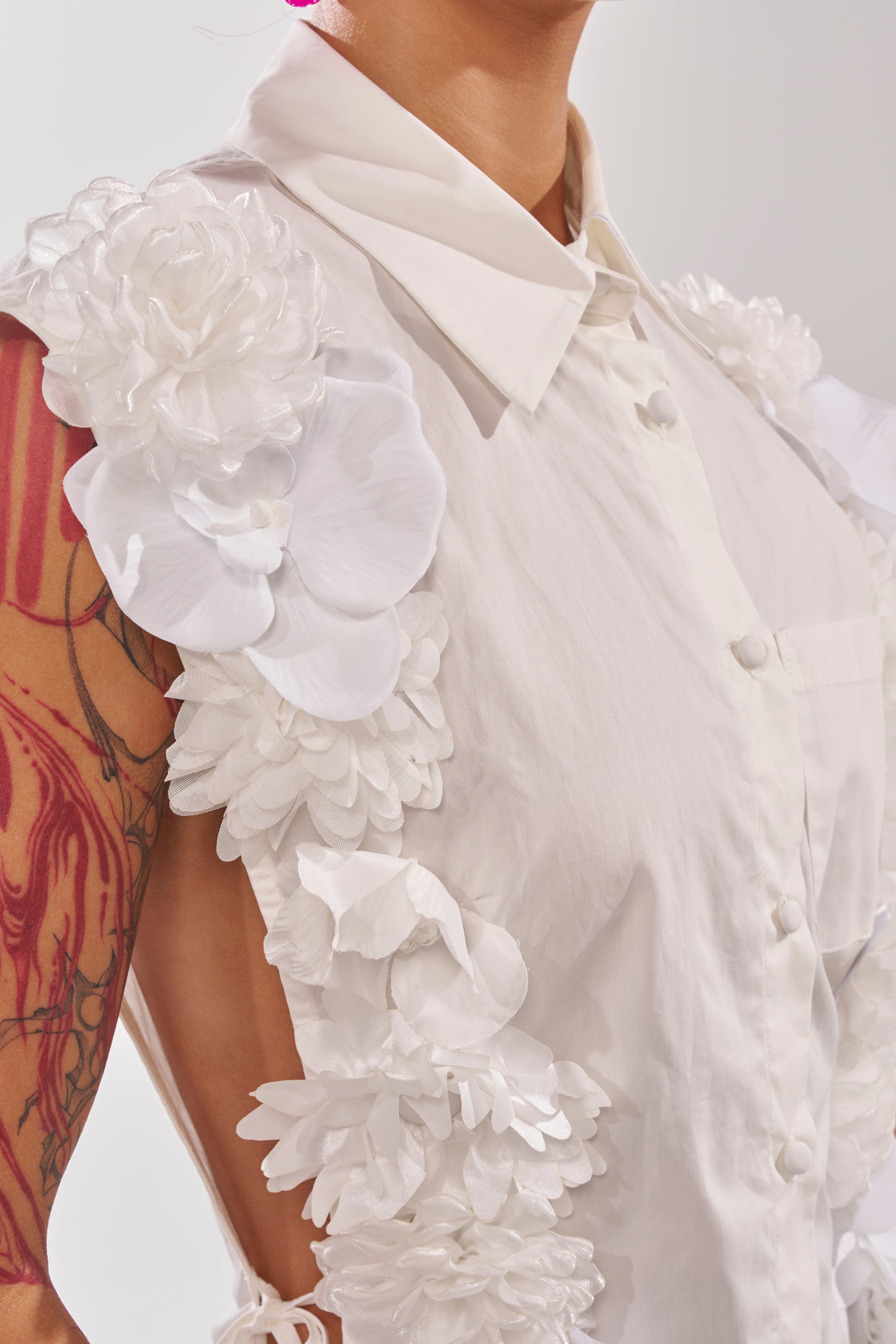 A person models the CRAZY FOR YOU BUTTON DOWN, a sleeveless white cotton blouse with bold 3D floral appliqué on the shoulders and sides, set against a softly lit plain background that highlights its unique design. 8