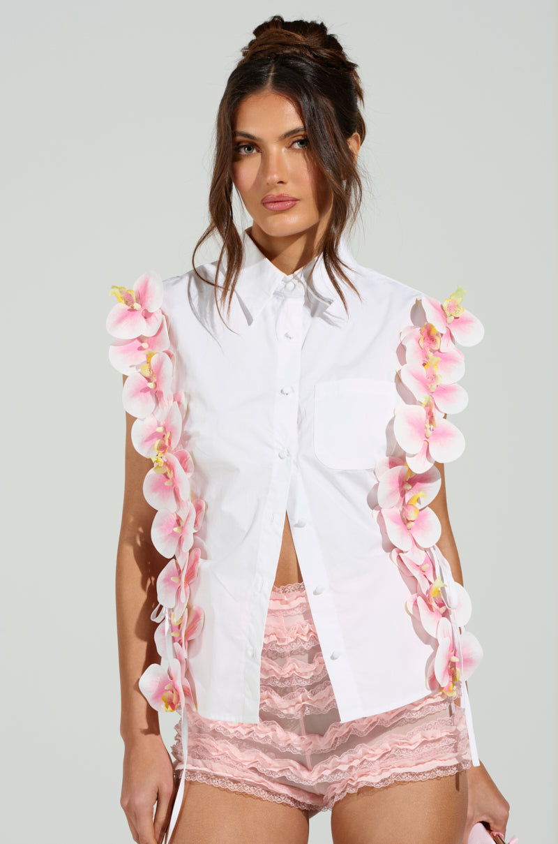 A woman wears a white, sleeveless FLOWER POWER BUTTON DOWN TOP featuring cascading pink florals paired with pink lace shorts. She stands against a light gray background with her dark hair styled in an elegant updo.