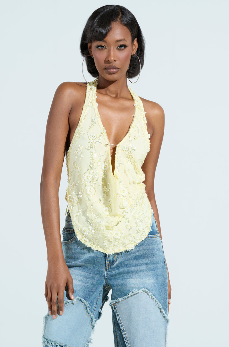 A person in a "Shine Bright Baby" yellow sequin halter top and blue jeans stands against a light backdrop, the top featuring floral appliques and a deep V-neckline. They have long dark hair and a neutral expression, perfectly capturing the vibrant vibe.