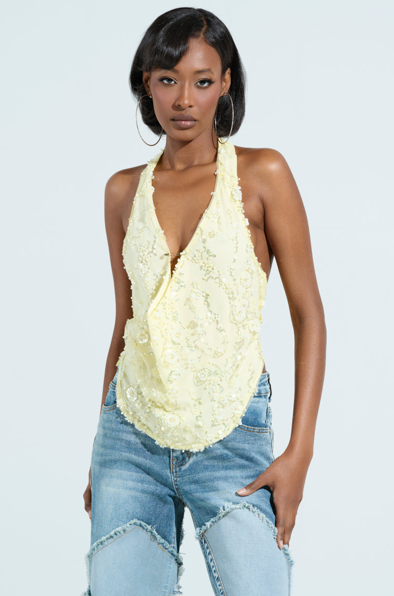 A woman exudes confidence in a SHINE BRIGHT BABY yellow sequin halter top with a deep V-neck and distressed blue jeans. Her long hair and large hoop earrings complete the look, as she poses with hands in pockets against a light gray backdrop. Shine bright, baby!.