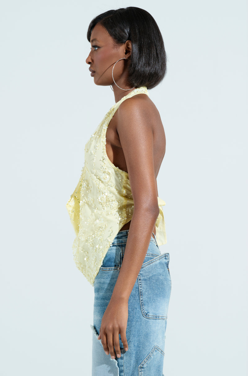 A person with short black hair is in profile, wearing the SHINE BRIGHT BABY YELLOW SEQUIN HALTER TOP with floral appliques and light blue jeans. They sport large hoop earrings against a plain white background.