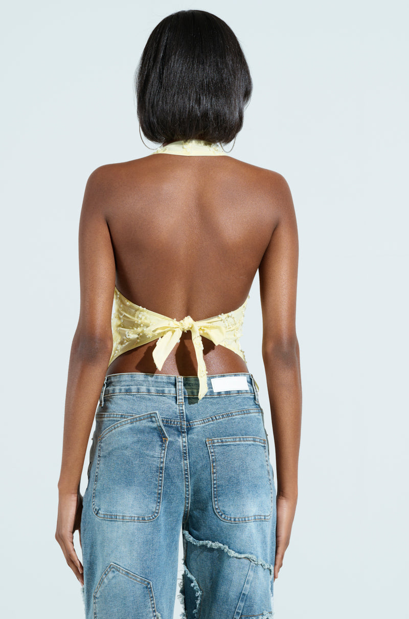 Rear view of a person with short dark hair, wearing the SHINE BRIGHT BABY YELLOW SEQUIN HALTER TOP tied at the back and distressed blue jeans against a light blue background.