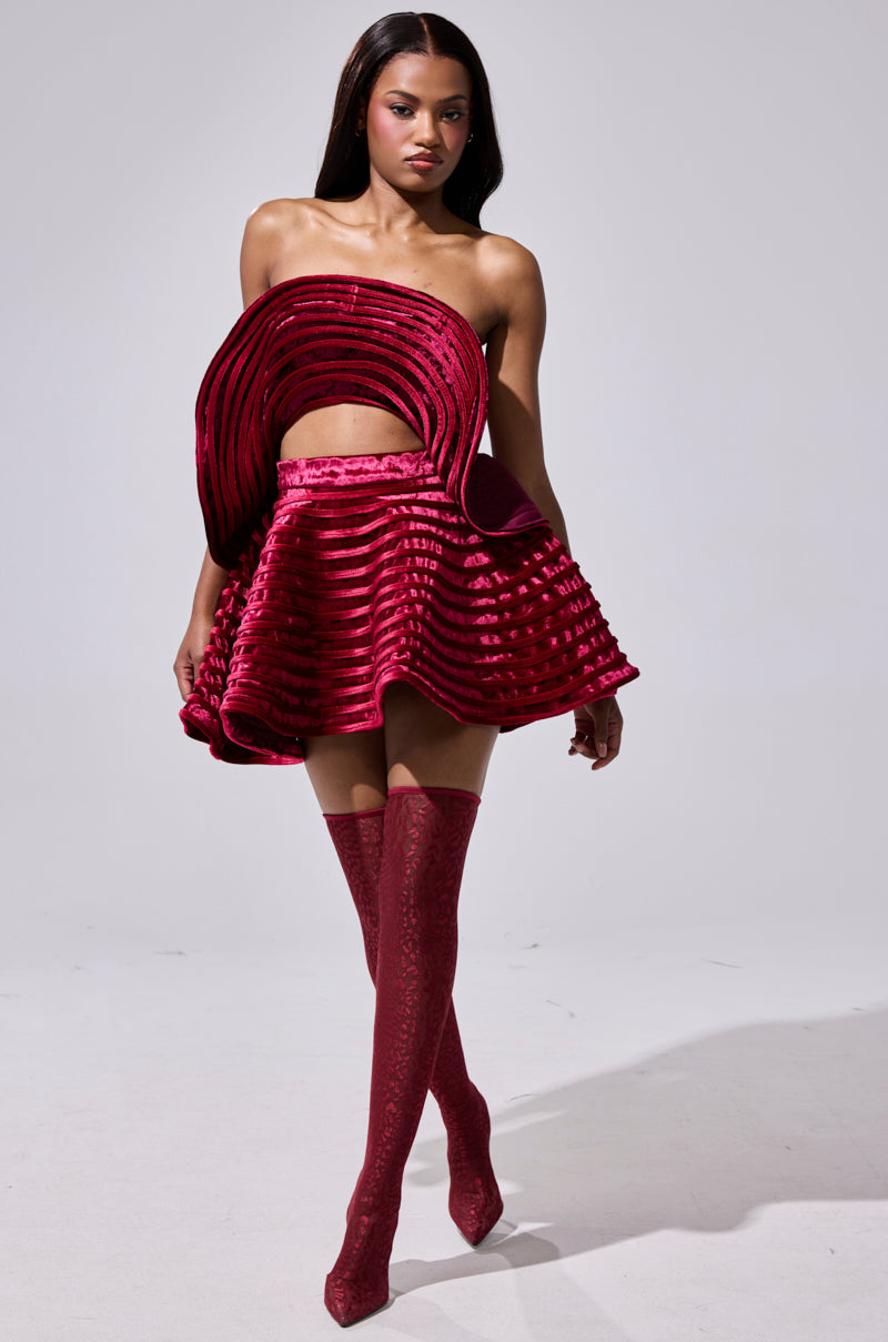 A woman wears the STAR OF THE SHOW VELVET MINI SKIRT in deep red, styled with matching over-the-knee boots against a plain gray background.