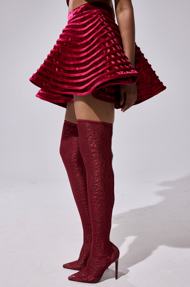 Wearing the STAR OF THE SHOW VELVET MINI SKIRT, a person pairs this structured, high-waisted red mini skirt with matching red patterned thigh-high heeled boots, standing against a plain light background.