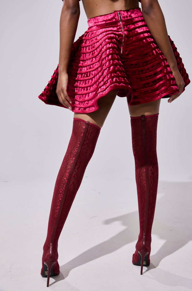 A person faces away, wearing the STAR OF THE SHOW VELVET MINI SKIRT—bright red, high-waisted with horizontal stripes—paired with thigh-high, heeled red boots. The background is plain and light-colored.