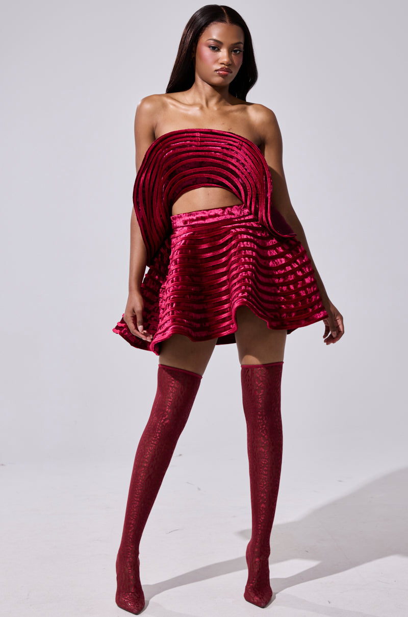 A woman models the STAR OF THE SHOW VELVET MINI SKIRT, a bold red velvet piece with circular ribbed layers and a high waist, paired with matching thigh-high boots against a plain light background.