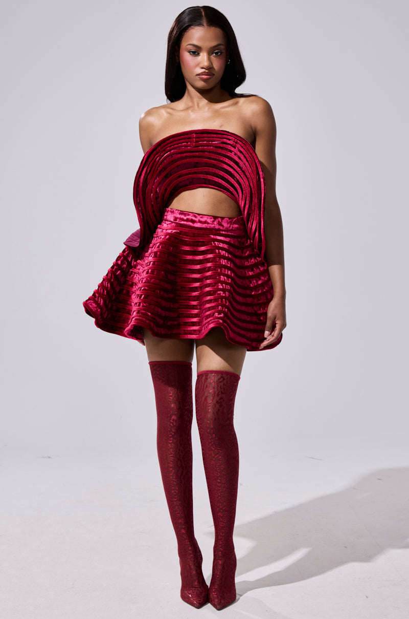 A woman wears the STAR OF THE SHOW VELVET MINI SKIRT in textured magenta with a high-waisted fit and layered circular patterns, paired with matching thigh-high boots against a plain light gray background.