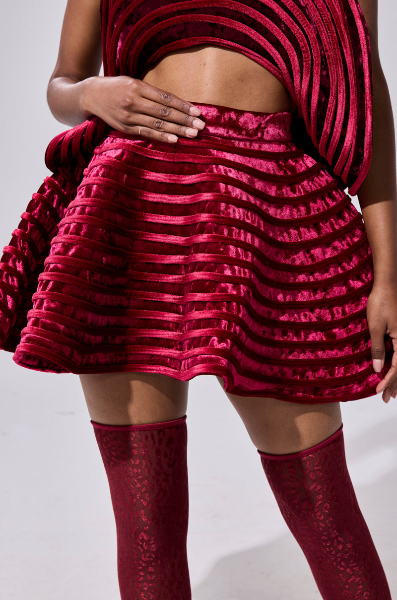 A person wears the STAR OF THE SHOW VELVET MINI SKIRT in deep red, featuring a textured, structured, flared shape with horizontal ridges. It's paired with matching thigh-high stockings and a sculptural top with layered and caged detailing.