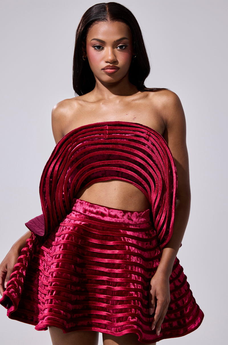 A woman poses against a neutral gray background wearing the STAR OF THE SHOW VELVET BLOUSE, featuring a vibrant deep red hue, textured velvet fabric, and sculptural, layered details for a striking, modern look.