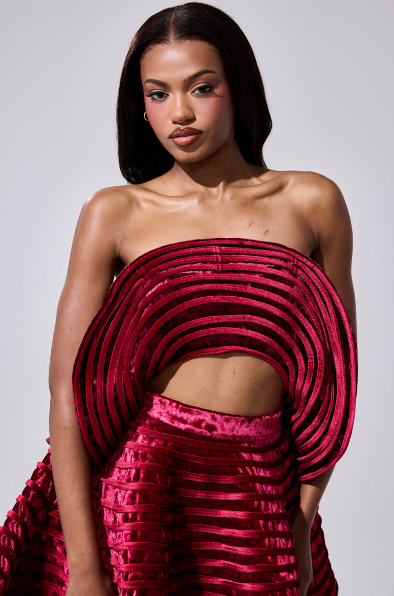 A woman wears the STAR OF THE SHOW VELVET BLOUSE, a striking textured deep red top with layered, curved lines and a ruffled overlay, standing against a plain light background and looking confidently at the camera.