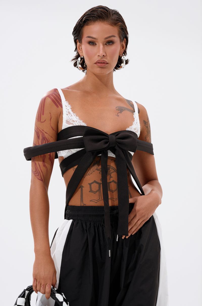 A person with short dark hair and visible tattoos wears the IM THE GIFT CHEST PIECE, a white lace bralette with an exaggerated bow, paired with black and white pants, standing against a plain white background. 0